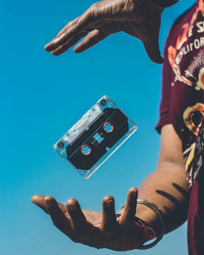 Home A retro cassette tape floating between hands against a clear blue sky, evoking nostalgia.